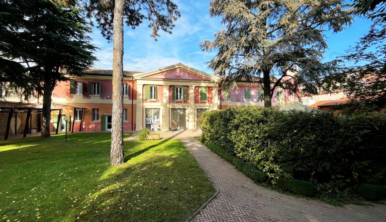 L'edificio Corte Bottacini, dalle tinte rosa, verdi e gialle. In primo piano ci sono alcuni alberi e un prato.