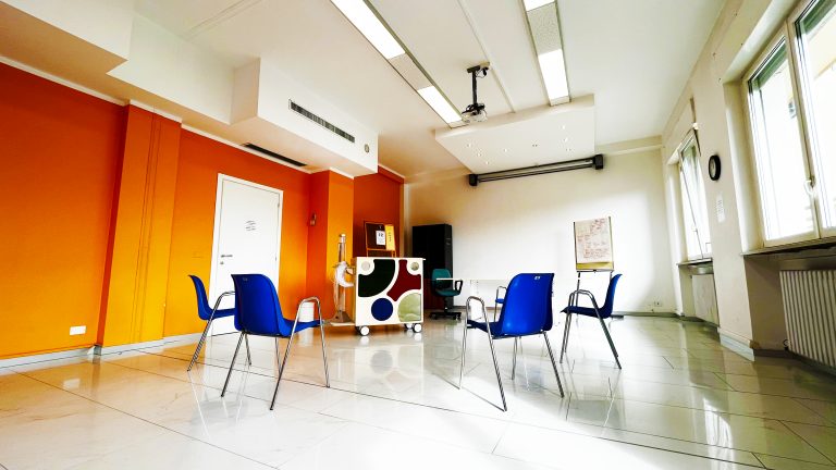 Nella luminosa sala, che mostra una parete arancione e un proiettore, alcune sedie sono disposte attorno al carrello Snoezelen.